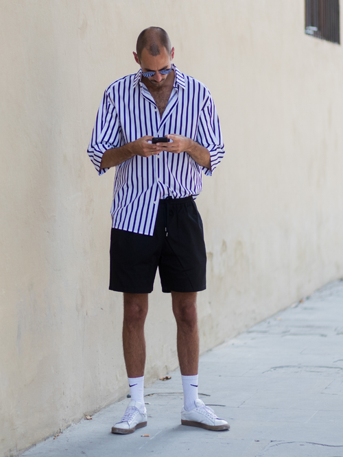 Street Style Look von der Pitti Uomo, wo ein Gast ein gestreiftes Hemd mit kurzer schwarzer Hose trägt.