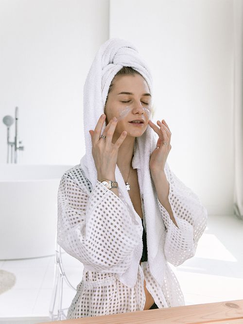 Frau im Bademantel und Handtuchturban trägt eine Gesichtsmaske auf.