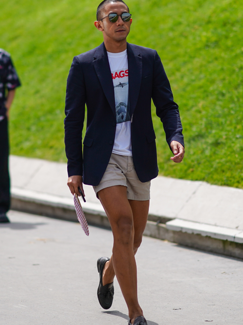 Street Style Look von der Paris Fashion Week, wo ein Gast ein dunkelblaues Sakko mit einem Print-T-Shirt und kurzer beigen Hose trägt.