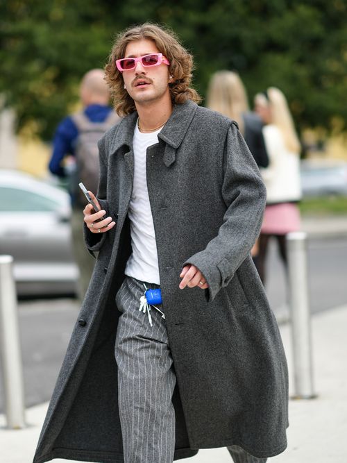 A guest wears pale pink sunglasses from Gucci, a gold chain necklace, a white ribbed pullover, a gray long coat, a royal blue AirPods case, gray with white stripes large pants, silver rings, white leather ankle boots, outside the MSGM fashion show during the Milan Fashion Week - Spring / Summer 2022 on September 25, 2021 in Milan, Italy.