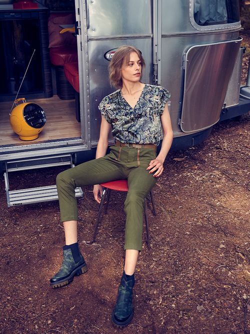 Frau in grüner Cargopants sitzt vor einem silbernen Wohnwagen und hört Radio.