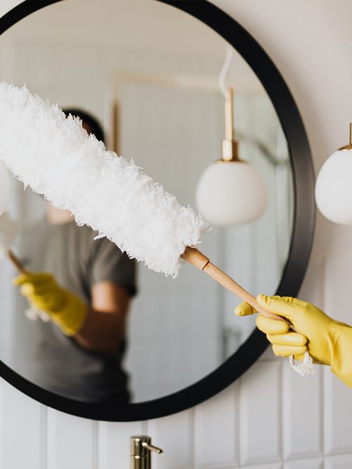 Wandspiegel wird mit einem weißen Staubwedel und gelben Gummihandschuhen gereinigt.