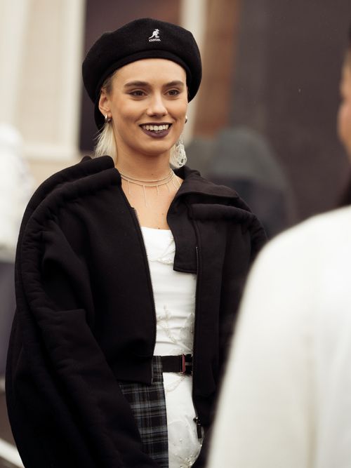 Street Style Look von der Visa Fashion Week, wo ein Gast einen schwarzen Mantel, weißes Shirt, Karorock und Kangol Mütze trägt.