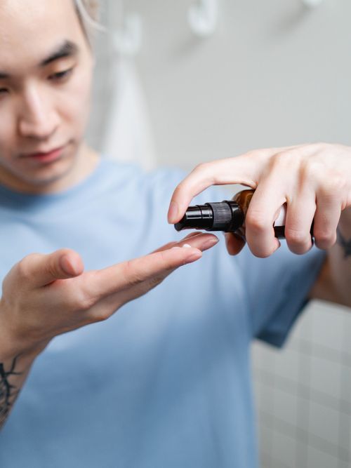 Ein Mann im hellblauen Shirt, der sich gerade im Badezimmer ein Serum auf die Hand aufträgt.