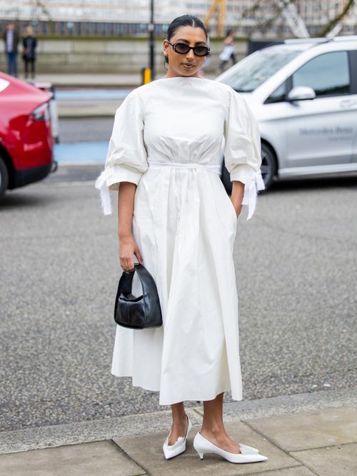 Street Style Look von der London Fashion Week, wo ein Gast ein weißes Wickelkleid mit Volant-Ärmeln, weiße Pumps und schwarze Tasche trägt. 