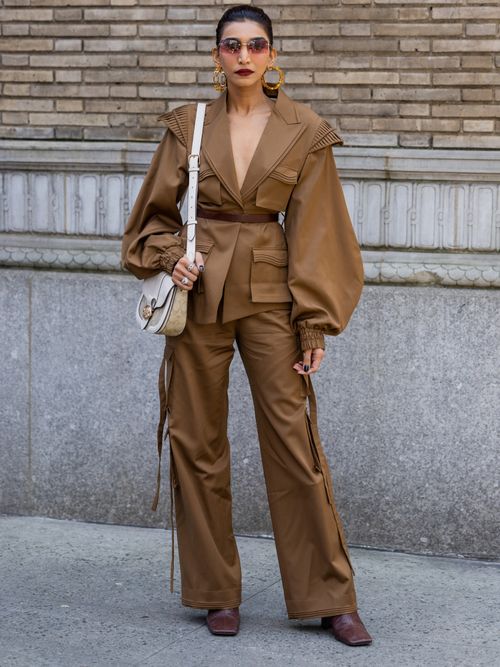 Juhi Godambe Jain trägt eine braune Jacke mit Taillengürtel und eine braune Hose. Dazu eine beige Tasche über die Schulter mit Sonnenbrillen und Creolen auf der New York Fashion Week.