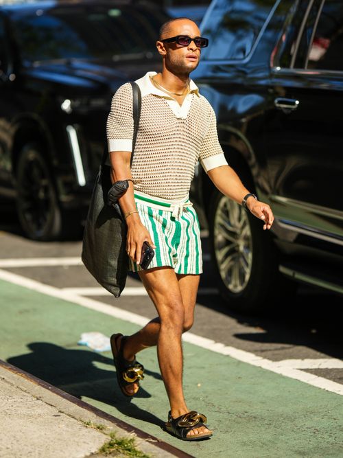 A guest wears black sunglasses, a white mesh / fishnet short sleeves polo shirt, a black wicker large shoulder bag, white latte and green striped print pattern shorts, a black shiny leather watch, black shiny leather with large gold metallic chain embroidered on the toe cap sandals , outside Altuzarra, during New York Fashion Week.