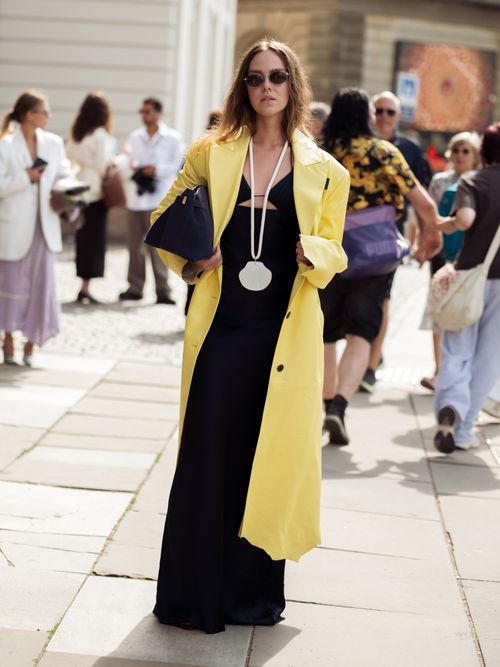 A guest wears black maxi dress. white shell statement necklace, yellow coat and navy blue bag outside the Mark Kenly Domino Tan show during day three of the Copenhagen Fashion Week (CPHFW) SS25 on August 07, 2024 in Copenhagen, Denmark.