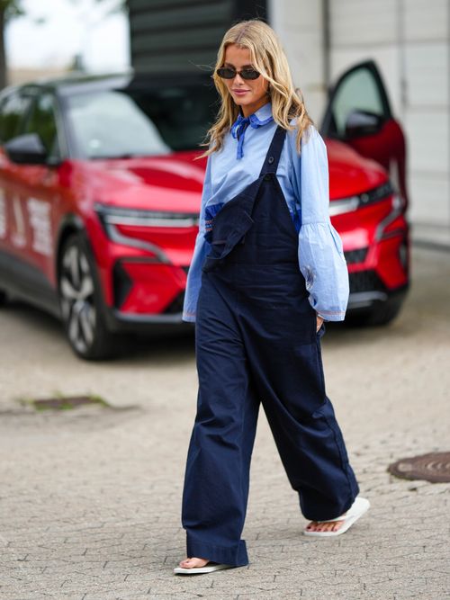 Street Style Look von der Copenhagen Fashion Week, wo ein Gast eine Latzhose mit blauer Bluse und weißen Flip Flops trägt.