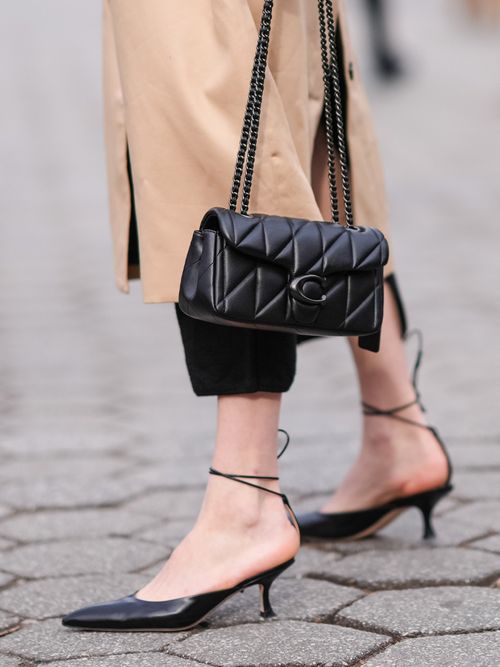 Close-up von einem schwarzen Kitten Heel mit Riemchen, schwarzer Crop-Hose, Trenchcoat und Coach Tasche.