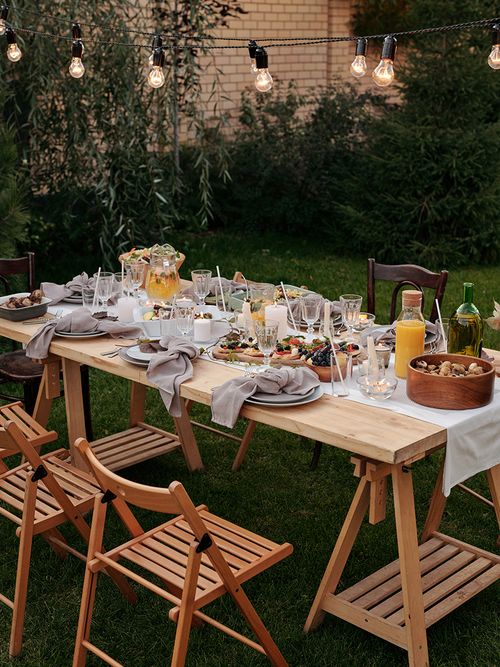 Reichlich gedeckter Tisch im Garten mit Holzstühlen und Lichterkette.