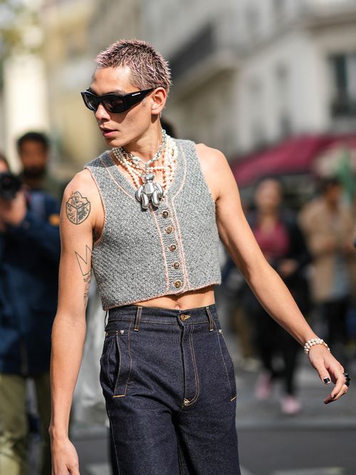 Evan Mock wears black sunglasses, a silver large chain large beard pendant necklace, a white large pearls necklace, a gray braided wool sleeveless buttoned copped t-shirt, pearls bracelets, navy blue high waist flared pants, outside Vivienne Westwood, during Paris Fashion Week - Womenswear Spring Summer 2022, on October 02, 2021 in Paris, France.