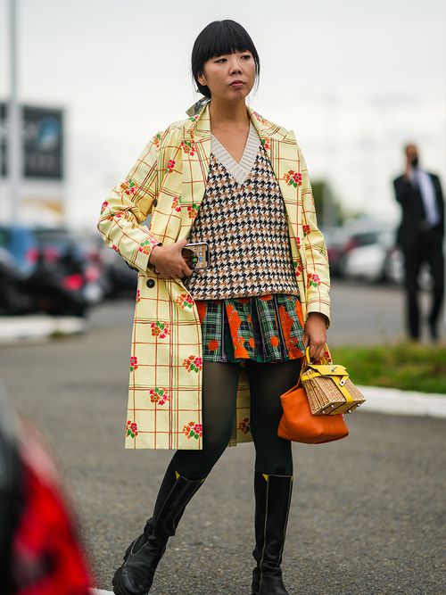 Susie Bubble in einem karierten Rock, einem Pullover mit V-Ausschnitt und einem gelb gemusterten Mantel.