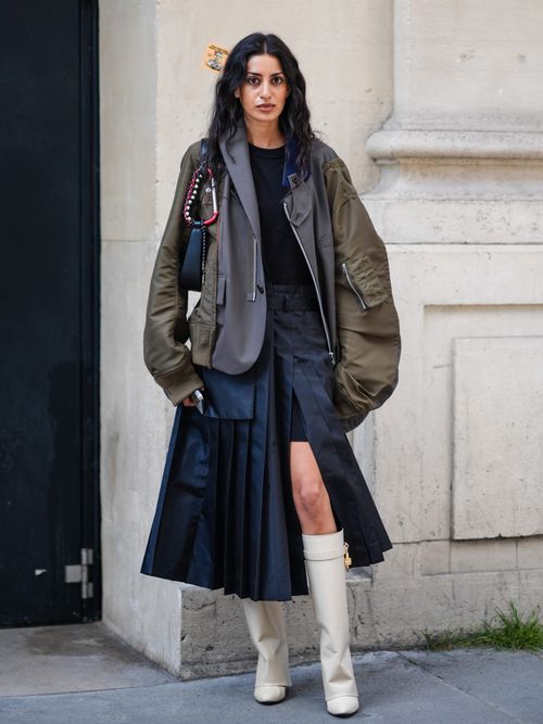 A guest wears black shirt, olive green and dark gray mix matched oversized bomber jacket, navy blue leather bag, navy blue pleated maxi skirt with high slit, white heeled boots Givenchy leather shoes, outside Sacai, during the Paris Fashion Week Menswear Spring/Summer 2025 on June 23, 2024 in Paris, France.