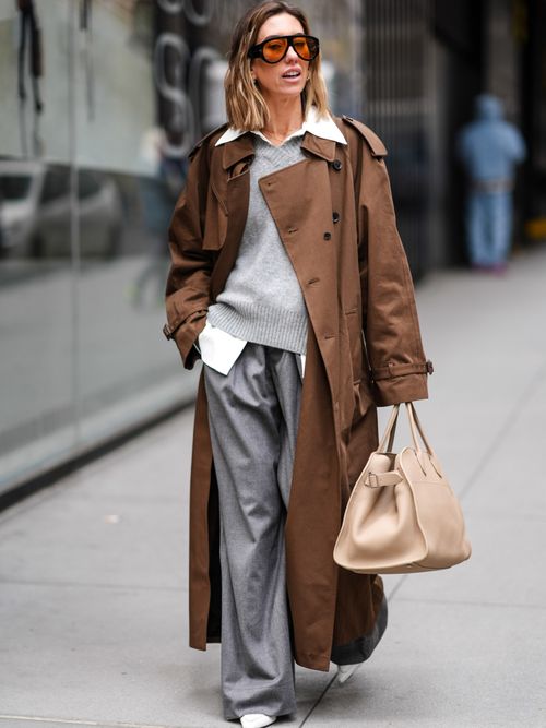 A guest wears sunglasses, a white shirt, a gray wool pullover , a brown trench long coat , gray flowing flared pants , white pointed shoes, a beige leather large bag, during New York Fashion Week, on February 12, 2024 in New York City.