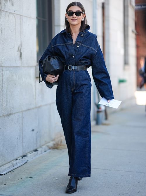Street Style Look von der Paris Fashion Week, wo ein Gast einen Denim-Jumpsuit mit schwarzen Gürtel, schwarze Bag, Boots und Sonnenbrillen trägt.