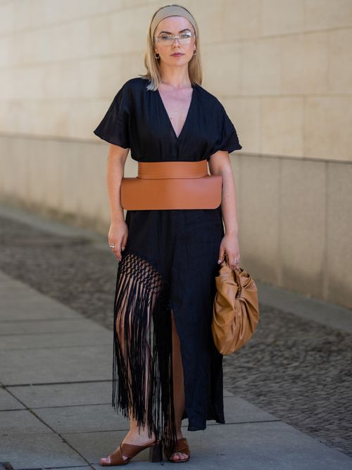 Amelie Stanescu wears a stretchy headband, frameless sunglasses, a black linen tunic dress, a tan leather waist belt, a tan leather shoulder bag, and tan leather mules.