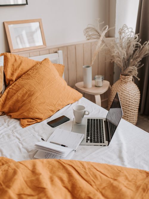 Bett mit orangefarbener Bettwäsche auf dem ein Laptop steht.