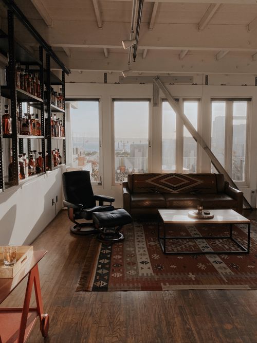 Interior shot featuring a black armchair and a brown couch with a patterned throw blanket in front of a large window. A metal bookcase with various bottles to the left and a bar cart.