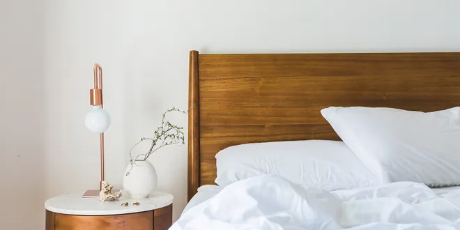 A wooden bedside table with a marble top. On it, a brass lamp and a white vase. Next to it, a wooden bed frame with white sheets.