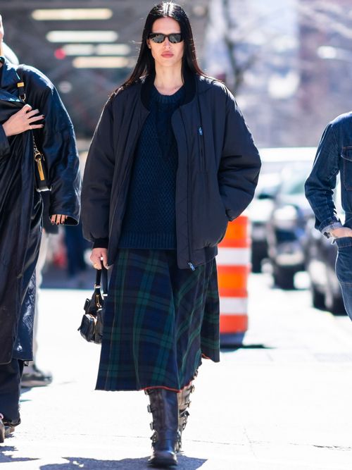Amelia Gray wears black sunglasses, a black bomber jacket, a black knitted jumper, a plaid pleated midi skirt, black boots, and a black leather bag.