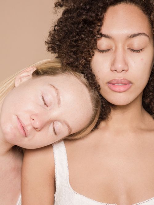 Blonde und dunkelhaarige Frau in weißen Tops und mit geschlossenen Augen.