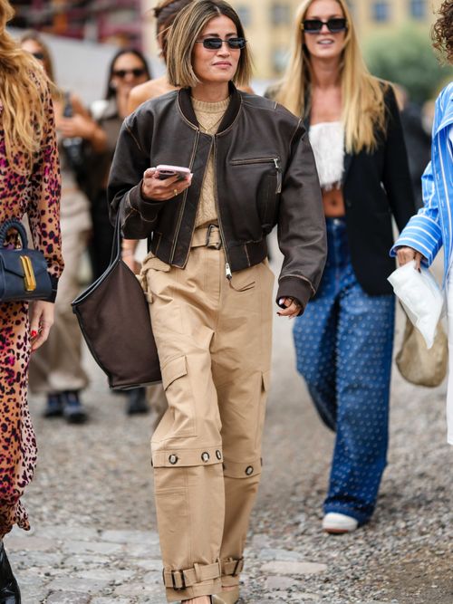 A guest wears black sunglasses, a beige braided wool pullover, a dark brown shiny leather zipper bomber coat, a brown handbag, beige belted cargo pants, beige shiny leather pointed pumps heels shoes , outside Munthe, during the Copenhagen Fashion Week Spring/Summer 2024 on August 10, 2023 in Copenhagen, Denmark.