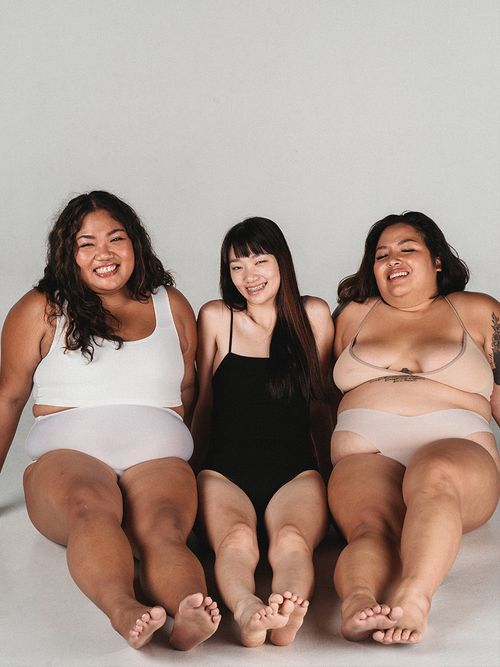 Drei Frauen in Unterwäsche die auf dem Boden sitzen und liegen und lachen.