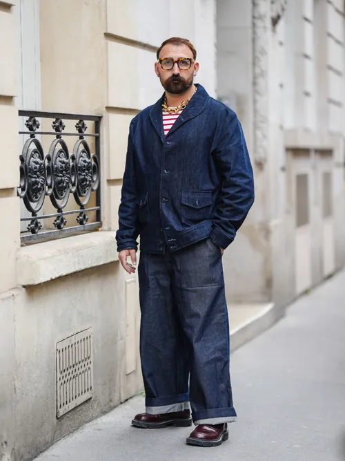 A guest wears brown sunglasses, silver earrings, a gold large chain necklace, a white and red striped print pattern t-shirt, a navy blue denim jacket, navy blue denim large pants, dark brown shiny leather shoes , outside Victoria Beckham, during Paris Fashion Week - Womenswear Spring/Summer 2023, on September 30, 2022 in Paris, France.