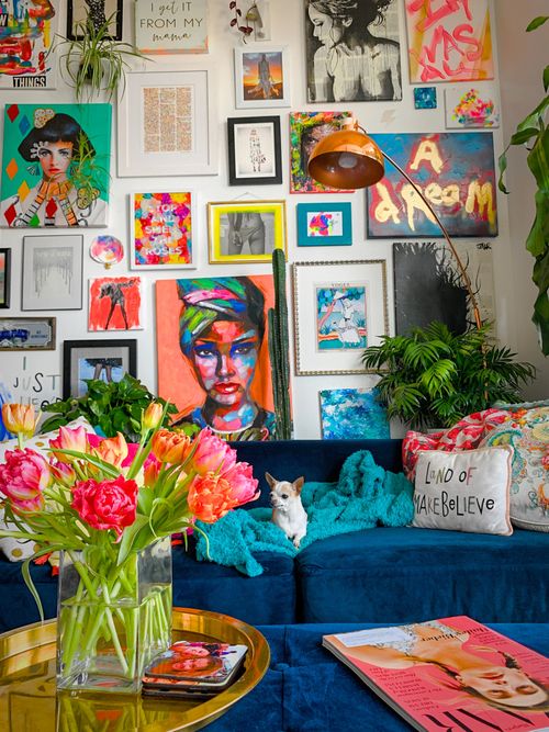 Buntes Wohnzimmer mit einer blauen Couch, vielen Bildern an der Wand und einem goldenen Couchtisch mit Blumen.