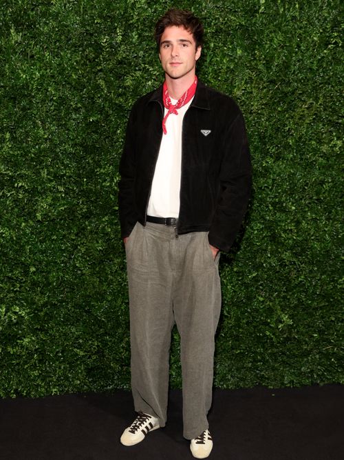 Jacob Elordi wears a red handkerchief, a white t-shirt, a navy worker jacket, grey slacks, and Adidas sneakers.
