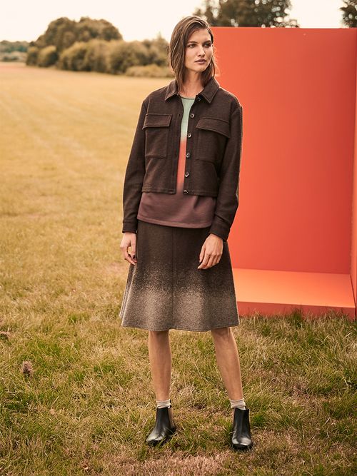 Frau mit Rock und brauner Jacke steht auf einer Wiese.