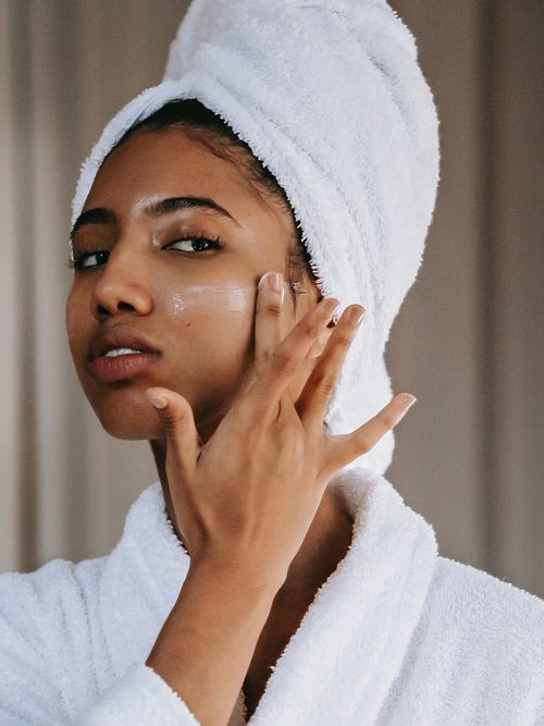 Frau mit Handtuchturban und Bademantel trägt eine Creme auf ihre Wange auf.
