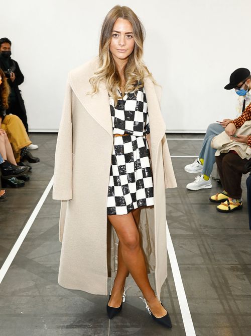 Frau mit einem beigen langen Mantel und einem Minirock und Top im Schachbrettmuster.