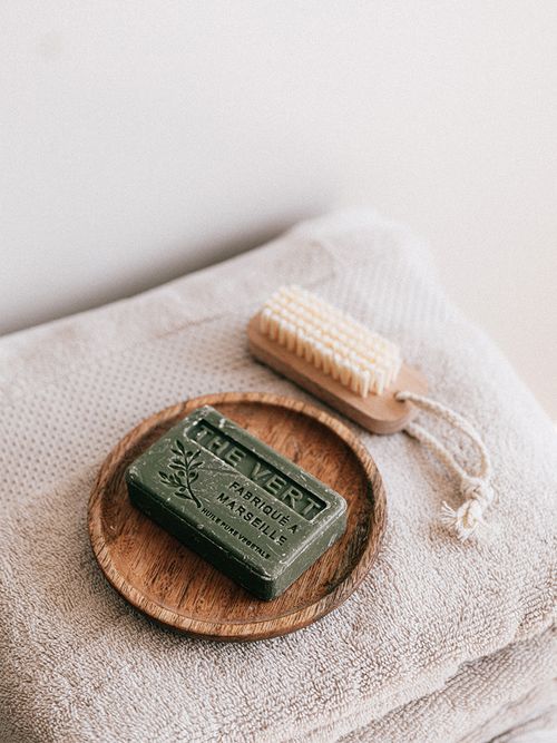 Beiges gefaltetes Handtuch auf dem ein Holztablett mit einer grünen Seife und eine Naturbürste liegt.