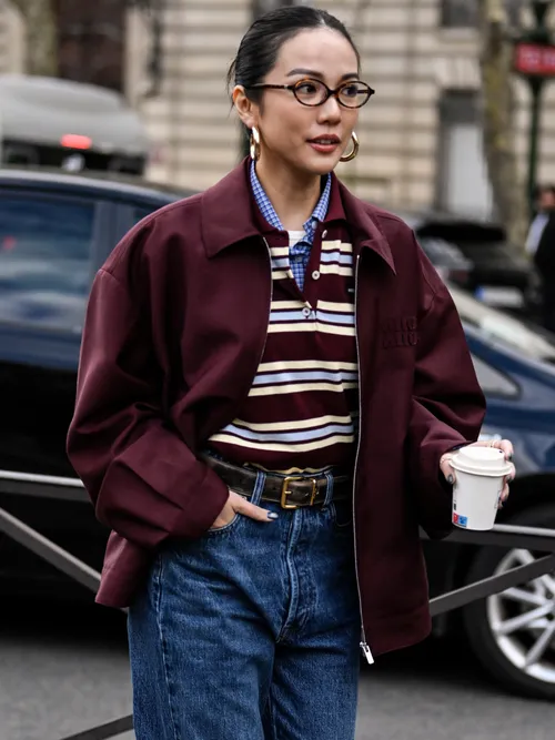 Yoyo Cao trägt eine bordeauxrote Jacke, Poloshirt, Halstuch, braunen Gürtel, Jeans und Creolen auf der Paris Fashion Week.