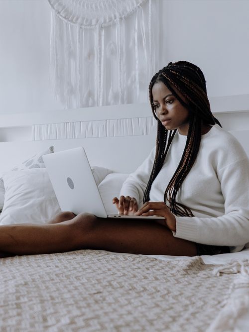 Frau sitzt auf ihrem weiß bezogenen Bett und schaut auf ihren Labtop.