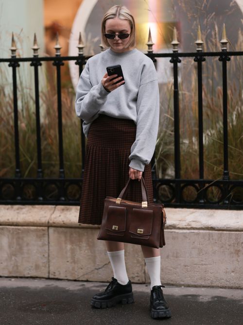 Street Style Look von der Paris Fashion Week, wo ein Gast einen grauen Pulli mit Faltenrock, Socken, Loafers und einer Aktentasche trägt.