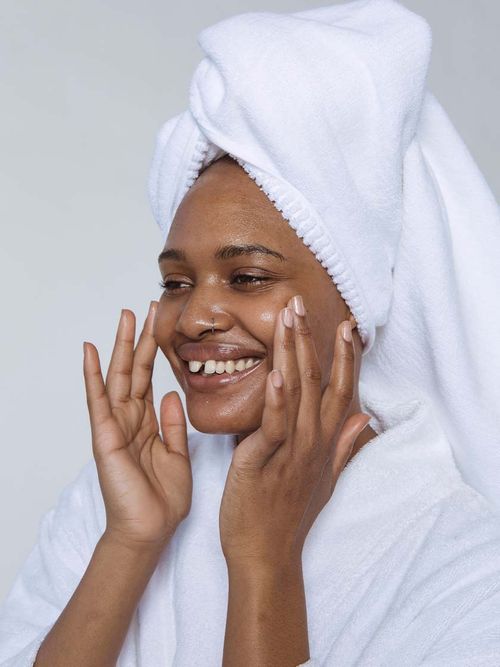 Frau in Bademantel und Handtuch auf dem Kopf schmiert sich lächelnd das Gesicht ein.