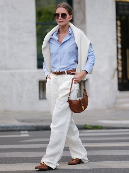 Diane Batoukina wears dark brown squared sunglasses, a pale blue cotton shirt, a white oversized pullover, a brown braided leather belt, baggy white linen pants, a camel brown handbag, and camel loafers.