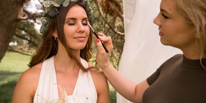 Braut mit Blumenkranz die gerade bei den Augen geschminkt wird