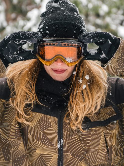 Portraitfoto von einer Frau auf der Piste und sie trägt eine schwarze Mütze, schwarze Handschuhe, Skibrille und eine braune Skijacke.