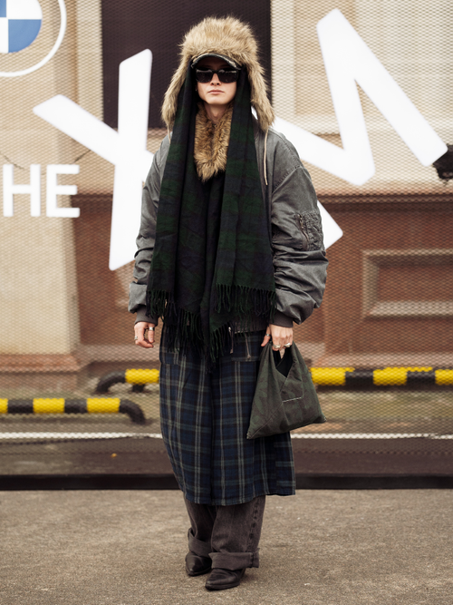 A guest wears black sweater, army green bomber jacket, dark brown jeans and green ceckered skirt, cap and fur hat and army green bag during the 10th season of the Visa Fashion Week Almaty on October 17, 2024 in Almaty, Kazakhstan.