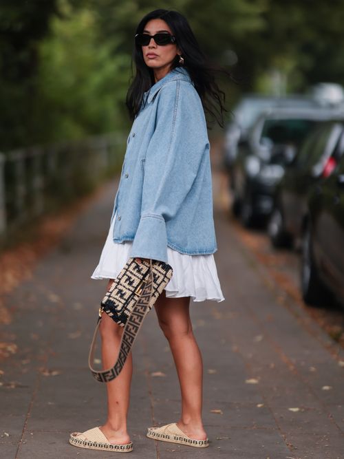 Semra Hunt wears a light wash denim jacket, a black and white Fendi monogram baguette bag, a pleated mini white skirt, and raffia sandals.