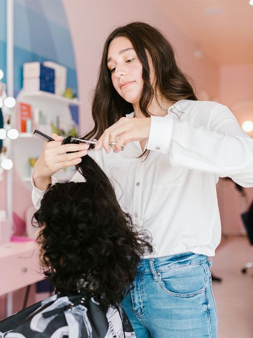 Frau in weißer Bluse und Jeans schneidet einer Frau mit Locken die Haare.