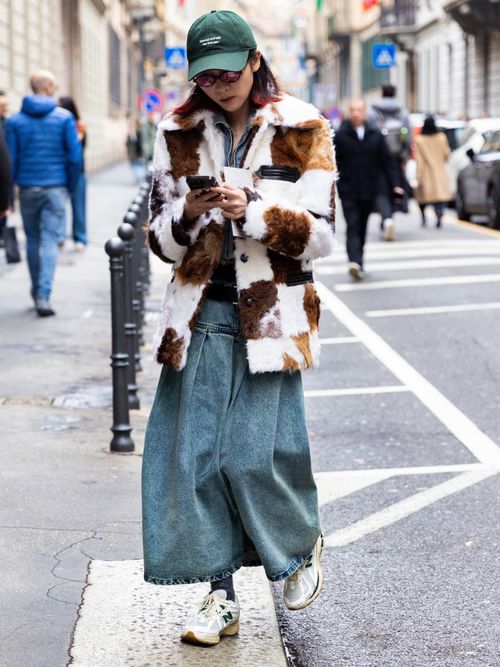 A guest is seen wearing a green hat with white littering details, a long denim skirt, white New Balance sneakers, grey lurex socks, a denim jacket and a white, brown and green fur jacket with black leather details outside Giorgio Armani show during the Milan Fashion Week - Womenswear Fall/Winter 2024-2025 on February 25, 2024 in Milan, Italy.