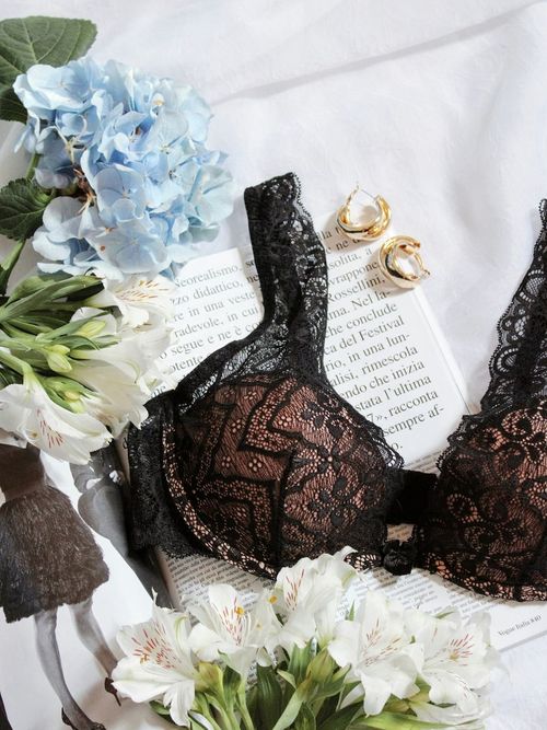 A black lace bra on a white sheet, an open magazine, gold hoop earringa, and blue and white flowers.