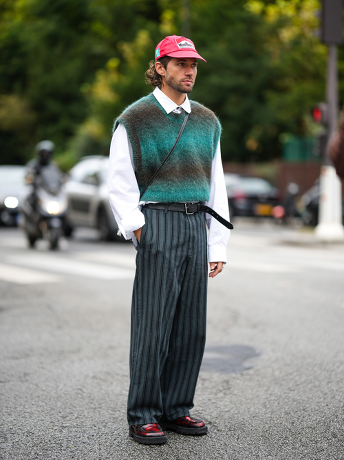 JS Rocques trägt einen trägt einen bunten Pullunder über ein weißes Hemd und Nadelstreifenhose mit roter Capy auf der Paris Fashion Week.