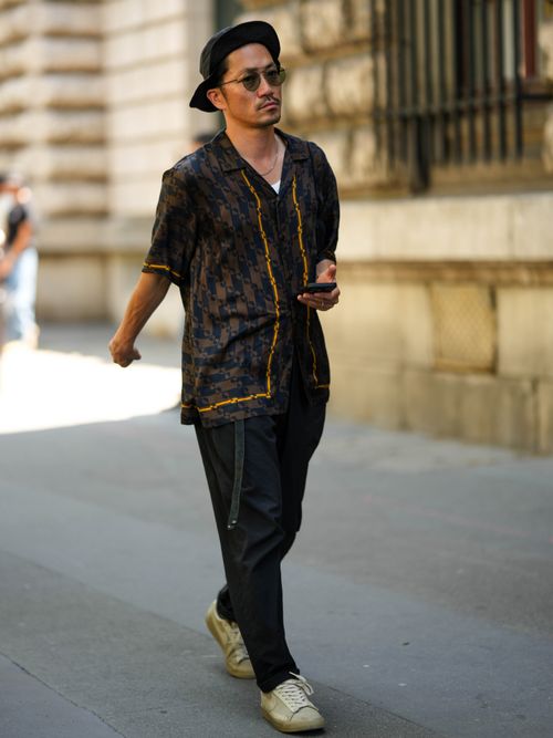 A guest wears a black felt wool hat, black sunglasses, a white tank-top, a dark brown and black print pattern with orange borders short sleeves silk shirt, black suit pants, beige suede and leather sneakers from Nike , outside Loewe, during the Menswear Spring/Summer 2024 as part of Paris Fashion Week on June 24, 2023 in Paris, France.