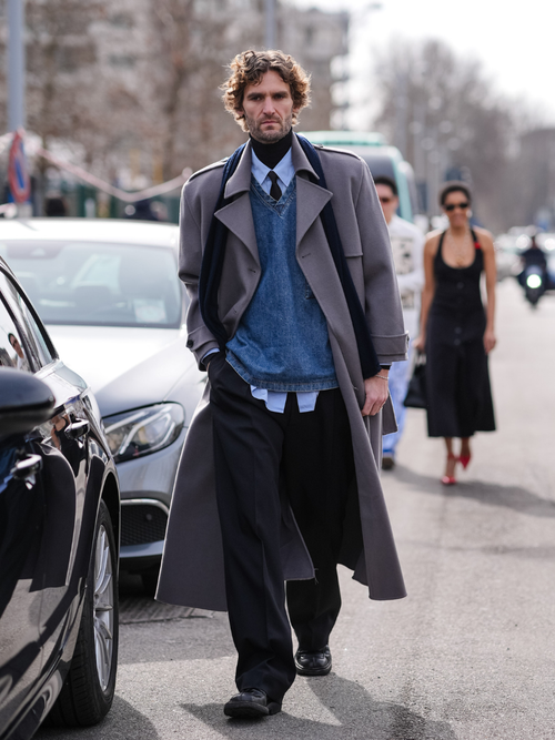 Street Style Look von der Milan Fashion Week, wo ein Gast einen grauen Trenchcoat mit V-Neck Pulli, Krawatte, Hemd und Rollkragen mit Jeans trägt.